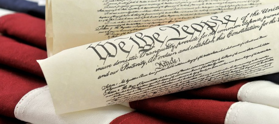 Rolled parchment of the U.S. Constitution displaying the words "We the People," resting on an American flag with visible red and white stripes.